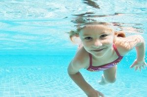 A young gril smilling and swimming underwater in a pool. Here are some helpful safety tips to keep you and your loved ones safe this summer and year-round around or in water.