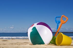 Day at the beach with a beach ball, spade and bucket in the foreground. May is National Water Safety Month.