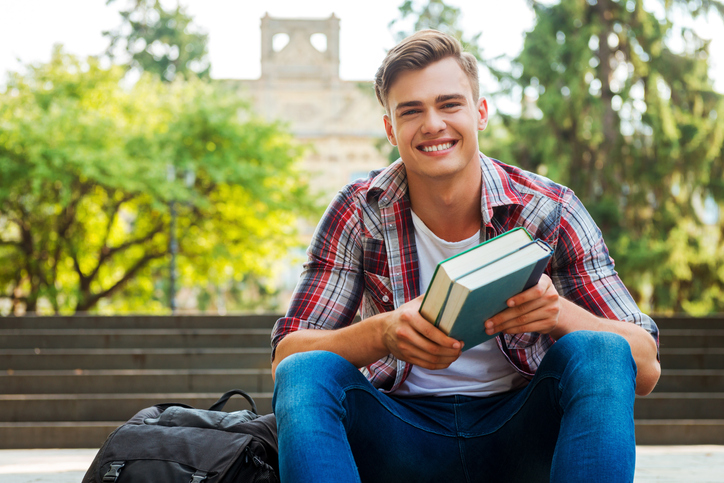 male student holding textbooks and smiling while sitting at the outdoors staircase with university building in the background