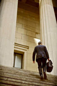 A Lawyer climbing steps framed by gigantic gothic pillars outside of a courthouse.